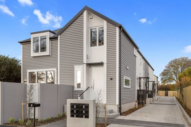 Exterior view of a narrow multi-storey townhouse or apartment-style residential building with grey horizontal weatherboard cladding, a steep gabled roof, white-trimmed windows and doors, and a long paved access path beside the building. The contemporary facade includes multiple levels and a shared mailbox structure at the front.