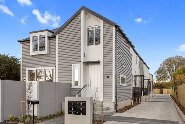 Exterior view of a contemporary multi-storey townhouse-style residential building with a steep gable roof, grey horizontal weatherboard cladding, white trim, and vertically arranged windows. A paved driveway runs alongside the building, with fencing, entry steps, and a shared mailbox feature visible in the foreground.