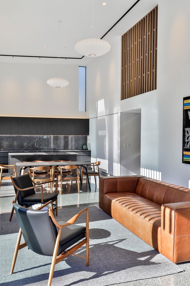 A bright, spacious contemporary open-plan living and dining area with a compact kitchen at the rear. The room features white walls and ceiling, dark timber kitchen cabinetry, a dark stone benchtop and splashback, a large dining table with timber chairs, a cognac leather sectional sofa, and mid-century-inspired leather lounge chairs. Vertical timber screening forms a feature wall high on the right, while sculptural pendant lights, concealed linear lighting, polished concrete or terrazzo-like flooring, and a large grey rug complete the refined interior. Strong sunlight creates defined shadows across the floor and furniture.