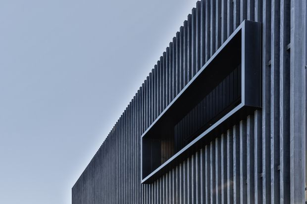 Close-up architectural exterior view of a dark, vertically ribbed facade with a projecting rectangular box-like window or balcony surround. The monochromatic composition emphasizes strong vertical repetition, sharp geometric edges, and deep shadowing against a pale sky.