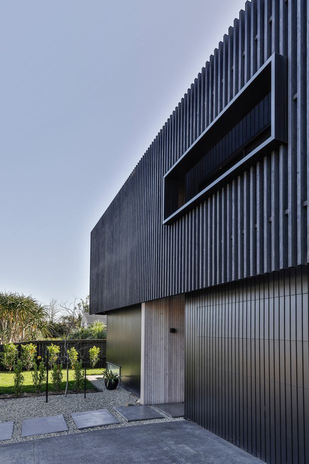 A contemporary two-storey residential facade viewed from the front driveway. The dark vertically ribbed exterior is paired with a projecting horizontal window surround and a tall black garage door. A pale timber-toned entry door, gravel landscaping, stepping stones, and restrained planting create a minimalist, highly geometric composition.