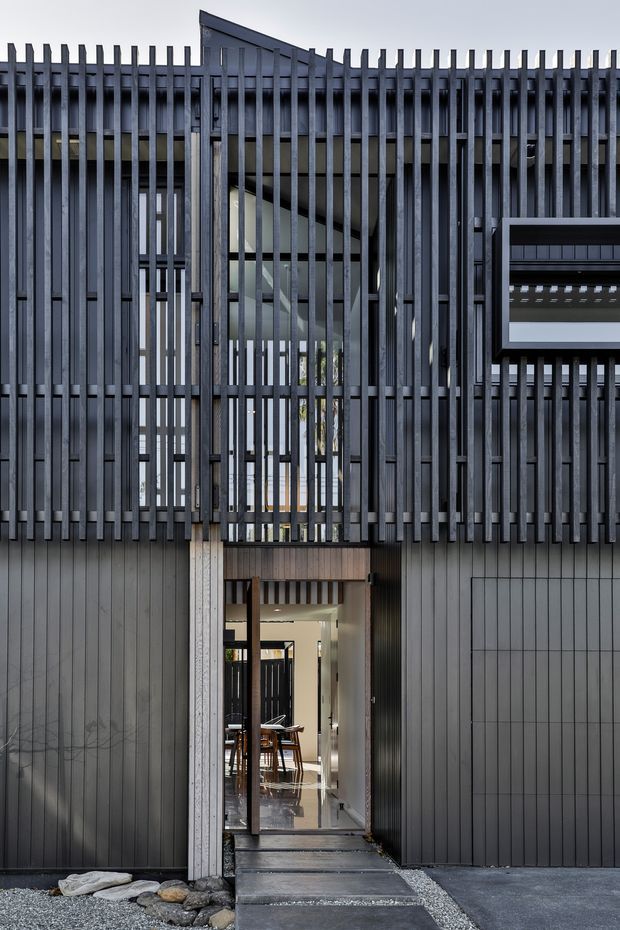 Front exterior view of a narrow contemporary multi-level residence. The facade combines dark vertical metal cladding and closely spaced vertical fins or battens, with a recessed glazed entry and a partially visible interior beyond. A sharply angled roof or upper volume rises behind the screen facade, creating a layered, privacy-focused composition. The palette is predominantly charcoal grey, black, and natural timber accents, with a gravel and stone threshold at the entrance.