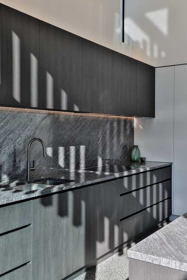 A contemporary minimalist kitchen with dark vertical-grain cabinetry, a dark polished stone benchtop and matching full-height splashback. The kitchen includes an undermount sink with a matte black gooseneck tap, integrated under-cabinet lighting, white wall cabinetry, and a grey stone island in the foreground. Strong sunlight creates graphic shadows across the dark joinery and stone surfaces.
