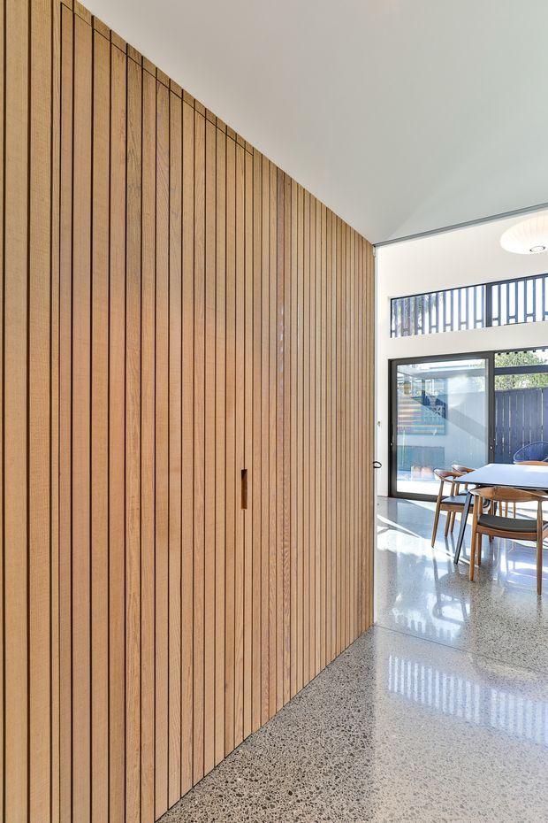 A bright contemporary interior featuring a full-height vertical timber slat wall with a concealed flush door. The wall curves or angles toward an open dining area with a timber dining table and chairs, large glazed doors, clerestory windows, and a polished exposed-aggregate concrete floor. The space has clean white ceilings, dark window frames, and strong natural daylight.