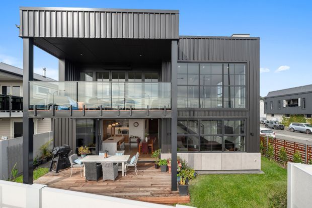 Contemporary multi-storey residential building viewed from the rear, featuring dark grey vertical metal cladding, extensive floor-to-ceiling glazing, a projecting upper-level volume, glass balustrades, and a timber outdoor deck with dining furniture. The image shows a compact landscaped yard and neighbouring urban residences.