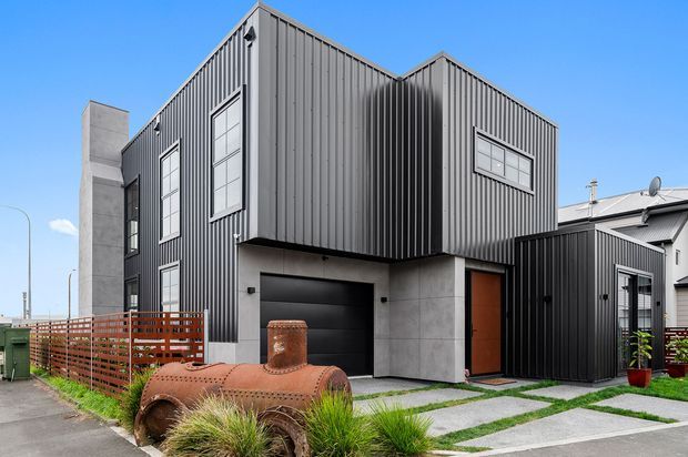Street-facing exterior view of a contemporary multi-level home with a sharply articulated gabled roof form, dark vertical corrugated metal cladding, concrete wall panels, and large black-framed windows. A recessed garage and timber-toned entry door sit beneath the projecting upper volume, with a slatted timber fence, concrete paths, lawn, and a sculptural rusted metal element in the foreground.