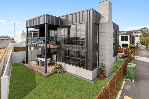 Exterior view of a contemporary multi-level residential home on a compact urban site. The house features dark vertical metal cladding, extensive black-framed glazing, projecting box-like forms, a concrete chimney or feature wall, a raised balcony with glass balustrade, and a small landscaped lawn and outdoor terrace.