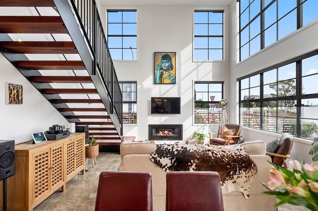 Bright, open-plan loft-style living room with a dramatic double-height volume, extensive black-framed industrial windows, and a floating staircase with dark timber treads and black metal balustrades. The room includes a large light sofa, cowhide throw, leather dining chairs, a timber lattice-front sideboard, fireplace, television, record player, artwork, and indoor plants. Strong natural light and exposed architectural openness create a contemporary industrial atmosphere.