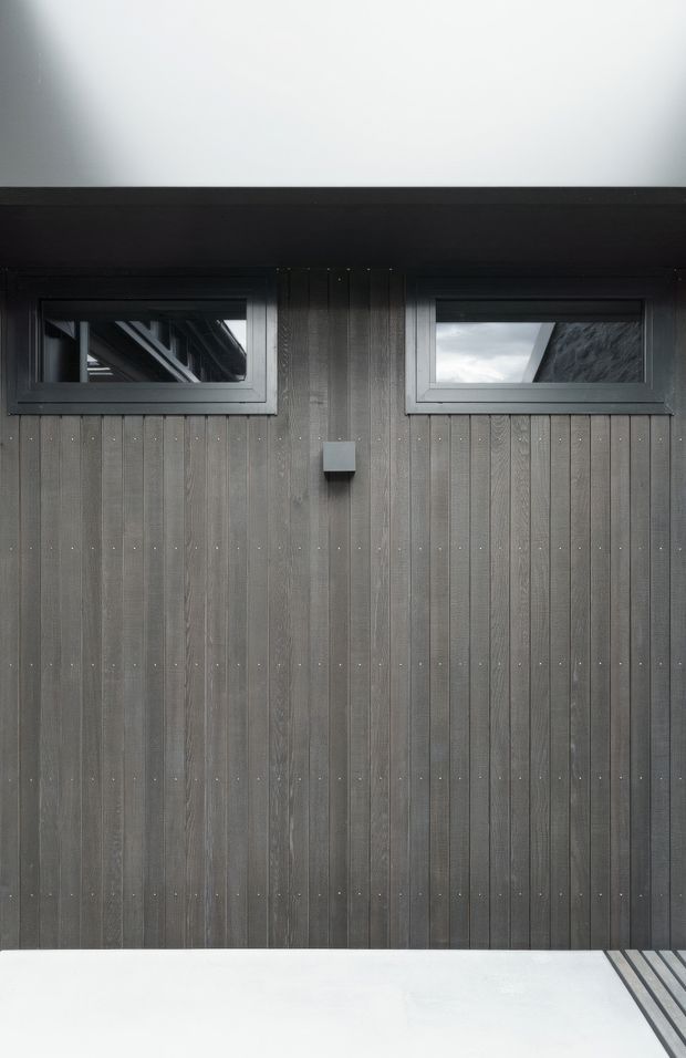 Close-up view of a dark, vertically timber-clad exterior wall with two narrow horizontal windows in dark frames and a small wall-mounted exterior light. A black roof or fascia band runs above, with a pale paved or decked surface in the foreground. The composition is minimalist and monochromatic.