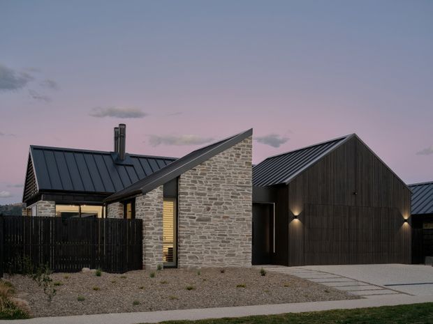 Exterior view of a contemporary single-storey residence composed of interconnected gabled volumes. The facade combines dark vertical timber cladding, pale irregular stone masonry, and dark standing-seam metal roofing. A gravel forecourt, concrete path, black fencing, recessed entry, and warm exterior wall lighting are visible at dusk.