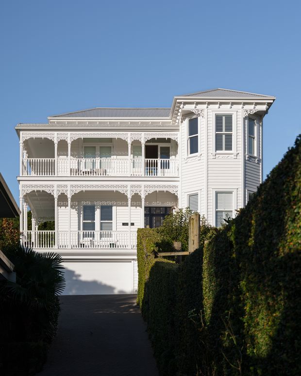 A stately two-storey white weatherboard residence viewed from a narrow driveway, featuring ornate lacework verandahs on both levels, decorative trims, shuttered windows, a projecting bay-like upper section, and a corrugated pitched roof. Tall clipped hedges frame the approach, with a clear blue sky beyond.