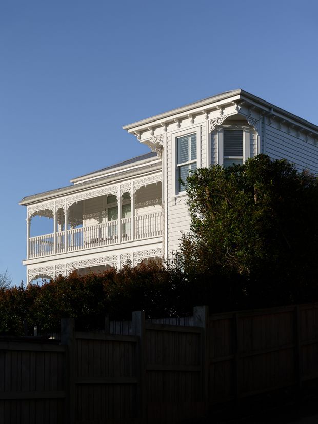 Exterior view of a historic ornate white timber house with multiple levels of decorative verandahs, intricate fretwork, balustrades, eaves brackets, and weatherboard cladding. The building is photographed against a clear blue sky, partially screened by dense hedges and a timber fence.
