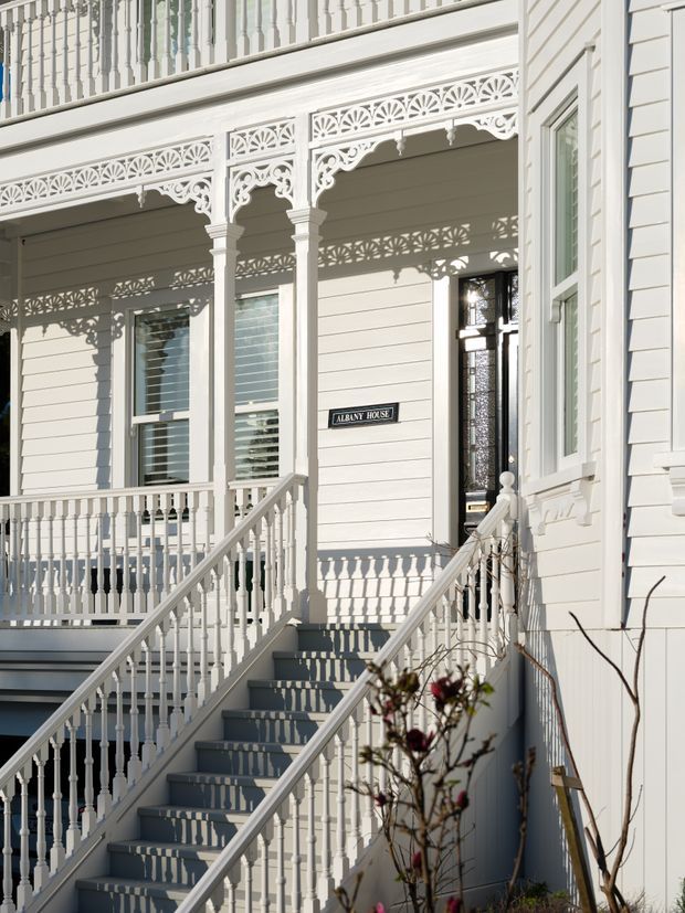 Exterior view of a highly detailed white-painted heritage townhouse or terrace facade, featuring ornate fretwork verandah balustrades, decorative brackets, turned posts, weatherboard cladding, sash windows, a stained-glass entry door, and a staircase leading to the raised entrance. A sign reads “Albany House.”