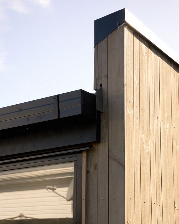 Close-up exterior architectural detail showing a modern timber-clad wall meeting a dark metal roof edge and garage door structure. The vertical light wood boards, crisp junctions, and strong sunlight emphasize the building envelope and construction detailing.