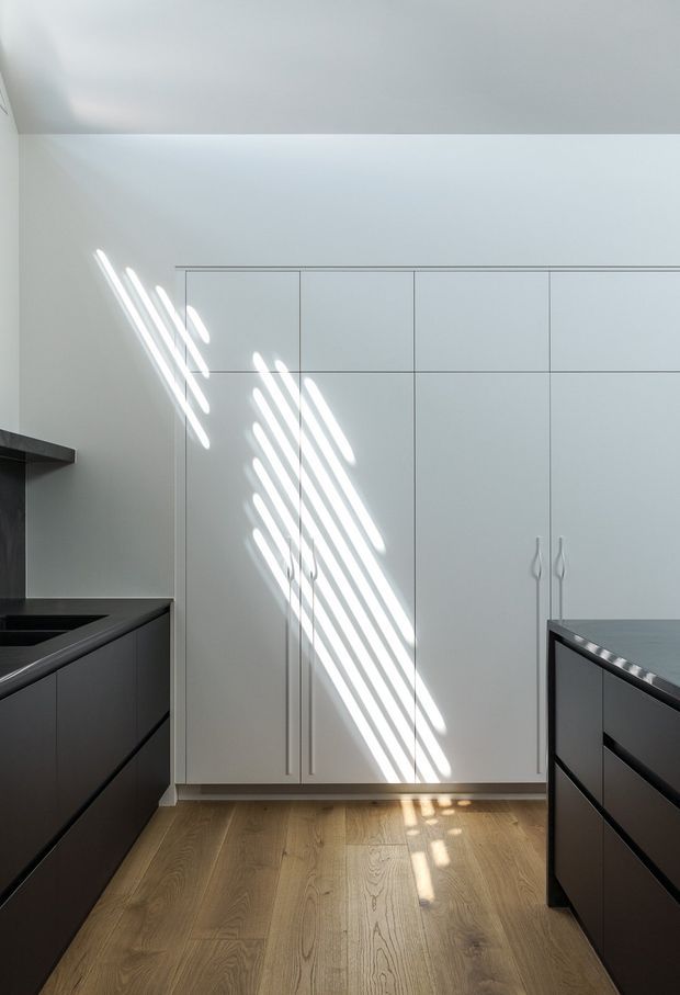 Minimal, light-filled contemporary kitchen with dark matte island cabinetry framing a bank of tall white flat-panel cabinets. Warm timber flooring contrasts with the white walls and cabinetry, while strong diagonal sunlight creates striped highlights across the cabinet fronts and floor.