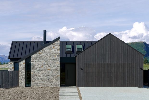 Exterior view of a contemporary, multi-volume house in a mountainous setting. The facade combines dark vertically ribbed cladding and standing-seam metal roofing with prominent rough natural-stone walls, a steep front gable, roof windows, and a broad pale concrete driveway.