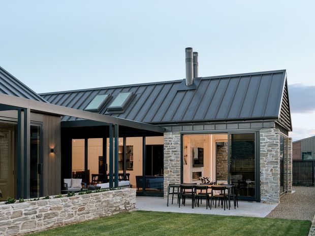 Exterior view of a contemporary single-storey home with a dark standing-seam pitched roof, extensive glazing, stone-clad walls, and a covered outdoor dining terrace opening onto a landscaped lawn and courtyard. The building has a connected wing form and visible roof skylights and chimney.