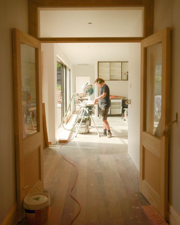 View through open timber-framed double doors into a bright space under renovation. A person is using workshop equipment beside large windows, with exposed or unfinished flooring, construction tools, cabinetry, and visible building work. The image is taken from an interior passage or doorway, with the work area beyond as the main visual focus.