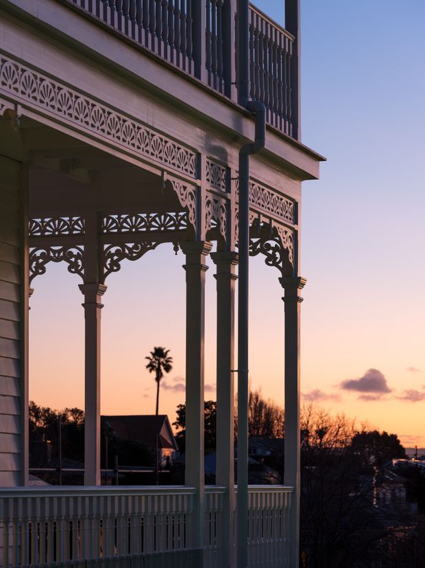 Close-up exterior view of a heritage-style verandah or balcony at sunset, featuring ornate fretwork, decorative timber columns, turned balustrades, and painted weatherboard cladding. The elevated structure overlooks a suburban landscape with silhouetted trees and rooftops.
