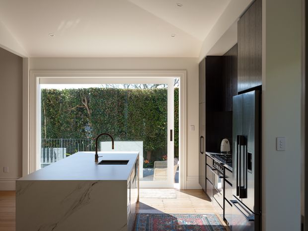 Bright contemporary kitchen viewed from the entry, featuring a large waterfall-edge island with an undermount sink and curved tap, dark full-height cabinetry with integrated stainless-steel appliances, pale timber flooring, and wide glazed doors opening to a leafy outdoor area. A patterned rug adds colour and texture.