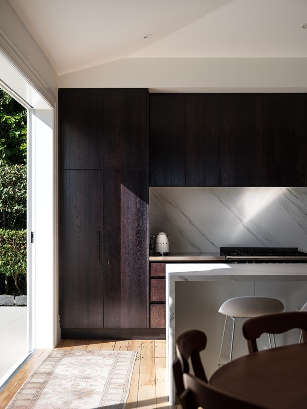 A compact, refined kitchen viewed from an adjacent dining area, featuring full-height dark timber cabinetry, a pale marble-look splashback, black cooktop, and a light island with integrated seating. Warm timber flooring, soft neutral walls, and large glazing create a bright contrast with the dark joinery. A dining table and chairs are visible in the foreground, and greenery is visible outside.