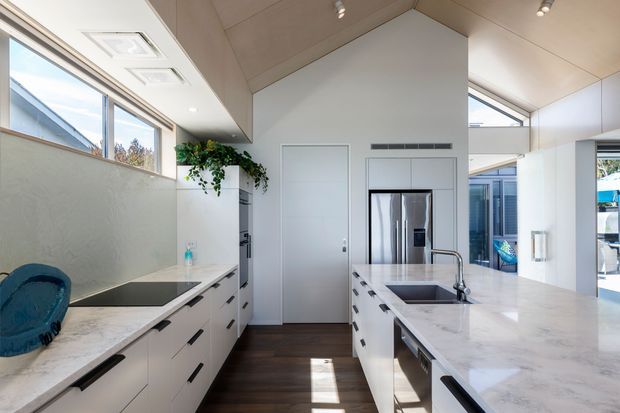 Bright contemporary kitchen with a central island, white flat-panel cabinetry, pale marble-look benchtops, dark timber flooring, stainless-steel appliances, and a high angled ceiling. Large clerestory and side windows provide abundant natural light, while a small indoor plant adds greenery.
