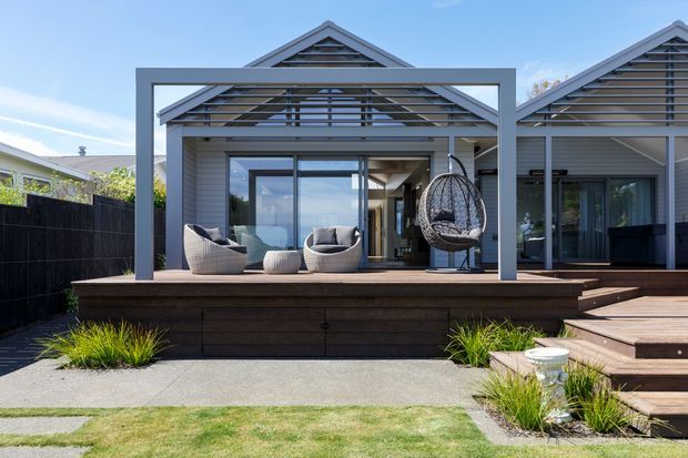 A contemporary single-storey home viewed from the rear garden, featuring a grey weatherboard facade, gabled roof forms, and a deep timber-framed covered outdoor terrace. The terrace includes outdoor lounge seating and a hanging chair, with broad timber steps leading to a landscaped lawn and paved path.