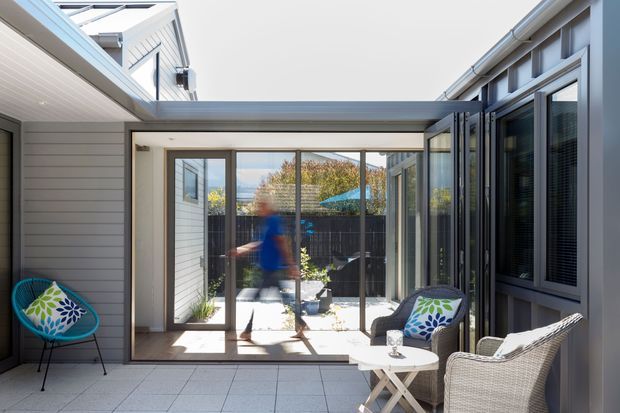 A bright enclosed courtyard or covered outdoor living area connecting residential wings. The space has pale tiled paving, grey horizontal cladding, extensive glass doors and windows, a glazed roof section, and an openable glass wall. Wicker-style seating, a small white folding table, and colourful accent chairs create a relaxed outdoor lounge atmosphere. A neighbouring or rear courtyard is visible beyond the glazing.