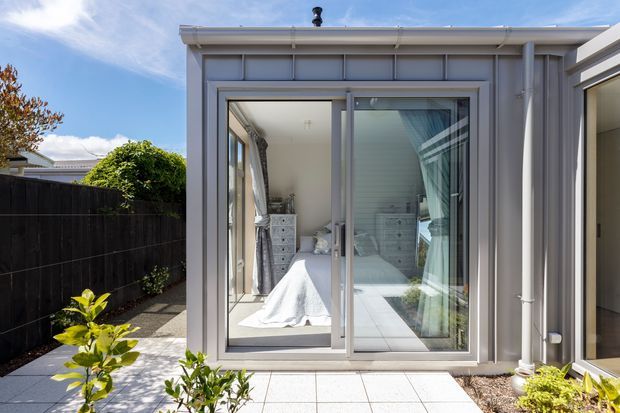 Exterior view of a compact contemporary bedroom extension with grey vertical cladding, a low-pitched flat roof, and large sliding glazed doors. The bedroom is visible through the glazing, with a bed, drawers, curtains, and bright white finishes. A paved patio and planted garden border sit in front of the extension, with a dark boundary fence to the side.