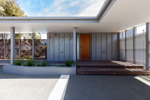 A bright contemporary single-storey residential entrance with a broad flat roof and covered porch. The facade combines pale grey vertical cladding, a warm timber front door, slim white posts, patterned privacy screens, and a dark timber entry deck. A raised planter with grasses separates the entry from the paved driveway, while vertical screening creates dappled shadows along the right side.