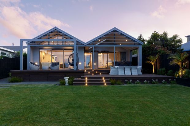 Twilight exterior view of a contemporary single-storey home with two prominent pitched gables and open timber-like slatted gable screens. The house opens onto a broad dark timber deck with outdoor seating, illuminated steps, landscaped lawn, and tropical planting. Large glazed openings connect the interior living areas to the deck.