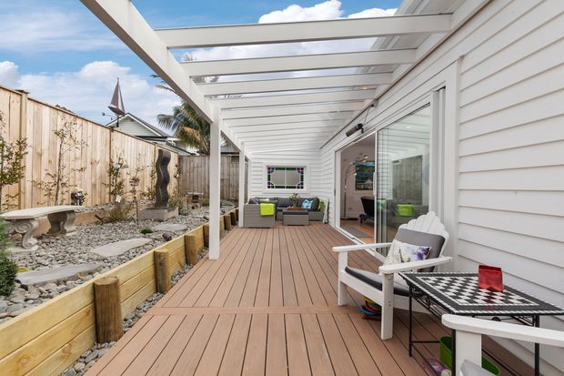 A bright outdoor deck and covered entertaining area alongside a white weatherboard-clad house. The space features timber decking, a white pergola with translucent roof panels, large sliding glass doors, outdoor lounge furniture, and a landscaped side garden with raised timber planters and decorative stone.