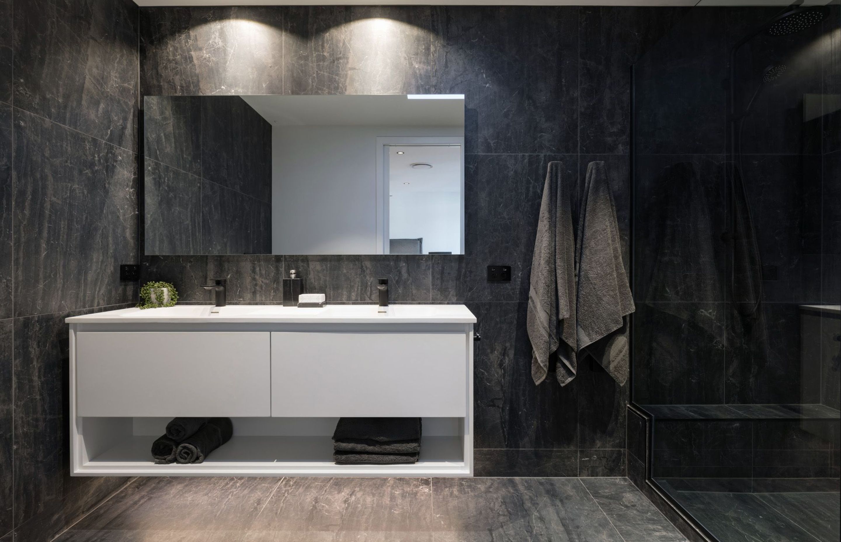 The crisp white bathroom cabinetry appears to hover within this dark space, which is clad in charcoal-grey tiles.