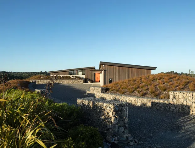 A low-profile modern building set within a landscaped, coastal-looking site. The structure has dark vertical timber cladding, broad low-pitched roof planes, extensive horizontal glazing, and a warm-toned entry door. Gabion stone retaining walls and gravel paths surround native-style planting, creating a strong connection to the rugged landscape.