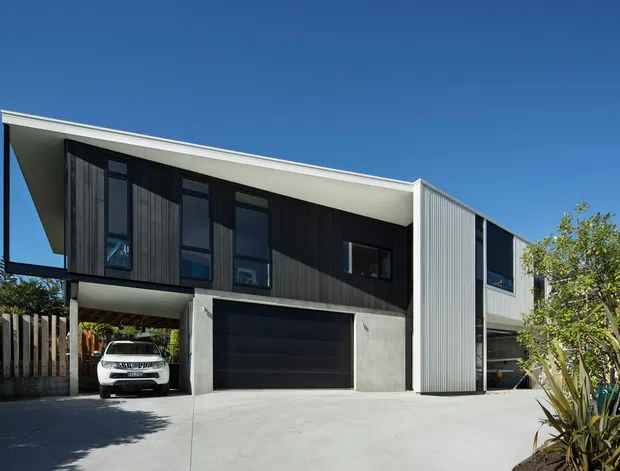 Exterior view of a contemporary multi-storey residence with a sharply angled roofline, dark vertical timber-look cladding, light corrugated metal cladding, concrete garage structure, and large dark-framed windows. A covered carport and integrated double garage open onto a broad concrete driveway, with established planting at the edge of the property.