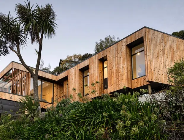 Exterior view of a contemporary, angular timber-clad house set within dense coastal-style landscaping. The building has a stepped, asymmetrical form with projecting volumes, deep window reveals, extensive glazing, and a partially screened upper-level terrace or balcony.