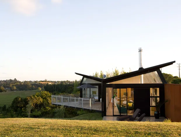 A contemporary single-storey house set in a rural landscape, photographed from the exterior at sunset. The compact dwelling has a distinctive asymmetrical folded roof with broad eaves, dark structural framing, extensive floor-to-ceiling glazing, and a raised deck or terrace with glass balustrades. A tall chimney and warm timber-toned wall section are visible, with open farmland and trees surrounding the property.