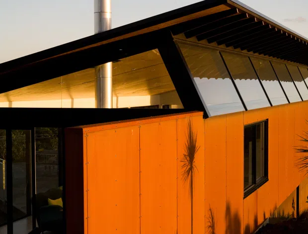 Exterior architectural photograph of a compact modern house at sunset, featuring vivid orange metal wall cladding, black steel framing, a sharply angled roof with exposed dark rafters, extensive sloping glazing, and a prominent stainless-steel chimney. A dim interior living area is visible through the glazed entrance.