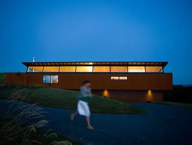 Twilight exterior view of a long, low modern house set into a landscaped sloping site. The building has a dark projecting roof with a continuous band of warm-lit clerestory glazing, brown timber-toned wall cladding, and a lower recessed level. A gravel driveway and planted grasses occupy the foreground, with a blurred person walking across the scene.