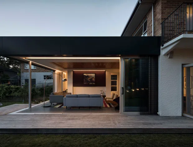 Contemporary residential exterior at dusk, showing a covered outdoor living extension opening onto a timber deck. The dark flat-roofed canopy contrasts with the warm timber ceiling, exposed brick upper level, white rendered walls, and extensive glazed doors. A furnished living room is visible through the open facade, with landscaped planting and neighbouring residential context in the background.