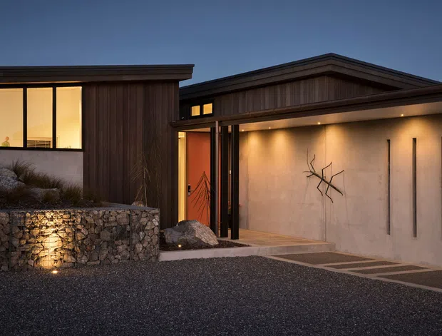 Twilight exterior view of a modern single-storey residence composed of low, rectilinear volumes. The facade combines dark vertical timber cladding with smooth rendered concrete-toned walls, extensive black-framed glazing, and a broad integrated garage or entry canopy. A rough stone retaining wall, gravel driveway, architectural lighting, and minimalist linear wall detailing define the approach.