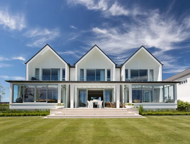 Front exterior view of a contemporary multi-level residential building with three prominent white gabled volumes, extensive floor-to-ceiling glazing, and a broad flat-roofed glazed lower terrace. The home opens onto a manicured lawn with low planting, under a bright blue sky.