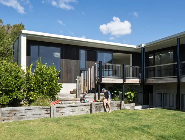 Exterior view of a contemporary multi-level residential house with dark vertical timber cladding, concrete elements, broad roof overhangs, and black metal-framed glazing and balustrades. Elevated decks and balconies overlook a landscaped lawn with timber retaining garden beds. The building is set against a leafy backdrop under a bright blue sky.