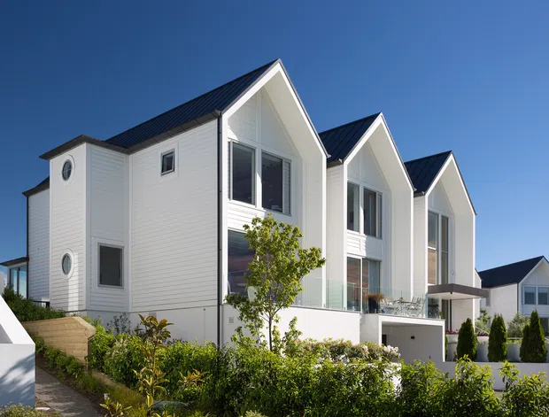 Exterior view of a row of contemporary multi-storey residences with steeply pitched dark roofs, white horizontal cladding, tall glazed openings, balconies, and integrated lower-level garages. Landscaped planting and paved access paths occupy the foreground under a clear blue sky.
