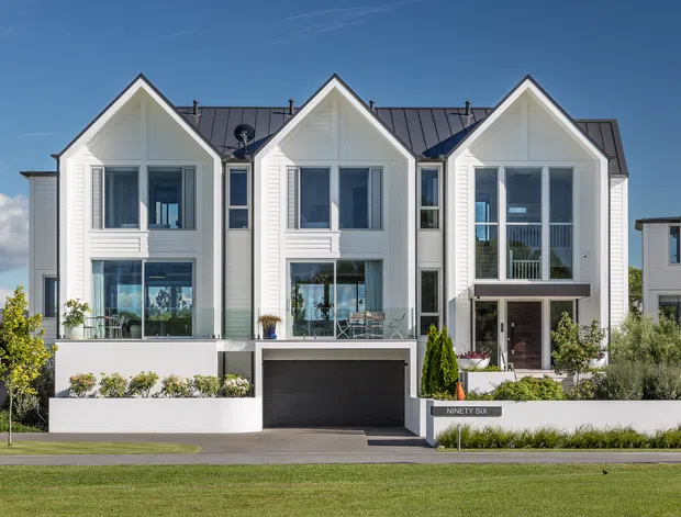 Front exterior view of a contemporary multi-storey townhouse complex with three prominent steep gabled bays, white horizontal cladding, dark standing-seam metal roofing, large vertically proportioned windows, balconies, and an integrated ground-level garage. Landscaped planting and low white boundary walls frame the street-facing facade.