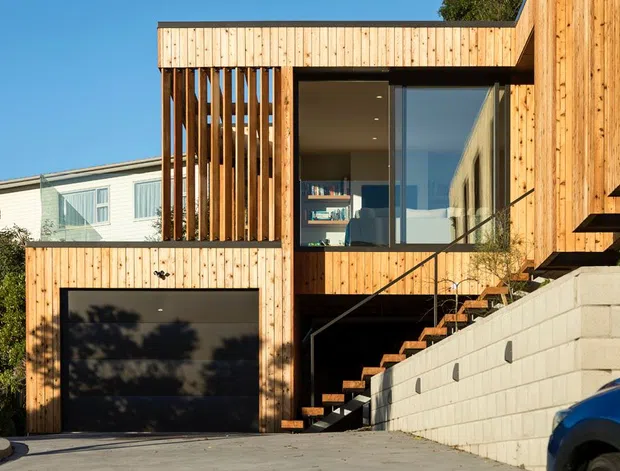 Exterior view of a contemporary multi-level residential house clad in warm vertical timber boards. The façade features a recessed dark garage, large glazed openings, a prominent external stair, vertical timber screening, and a pale concrete-block retaining wall on a sloped urban site.