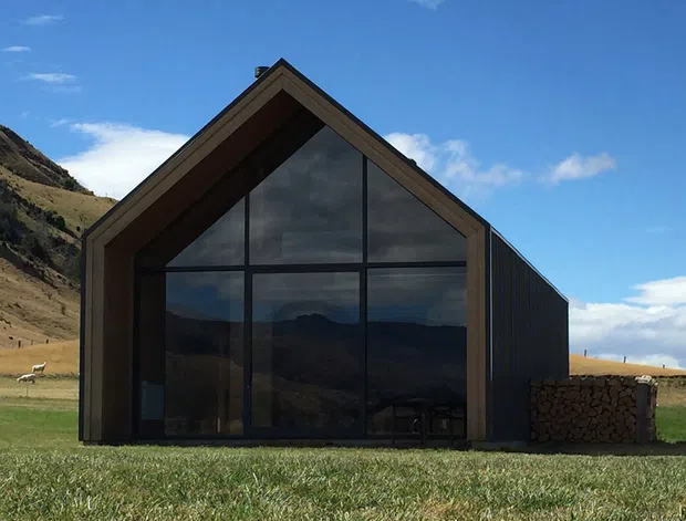 A small contemporary barn-like building set in an alpine rural landscape. The front facade has a steep gabled roof, dark timber framing and cladding, and a large floor-to-gable glazed entrance reflecting the surrounding mountains. A stacked wood feature sits beside the structure, with pasture and steep hills behind.
