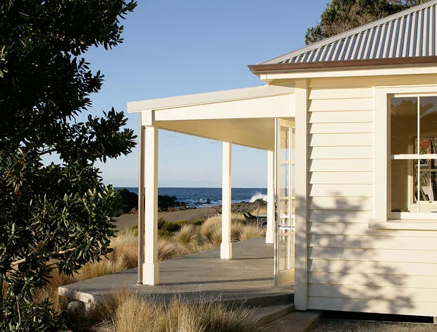 A coastal single-storey cottage exterior with cream horizontal weatherboard cladding, white trim, a covered open porch, and a corrugated metal hipped roof. The building overlooks a sandy shoreline and ocean, with native grasses and shrubs around a concrete path and terrace.