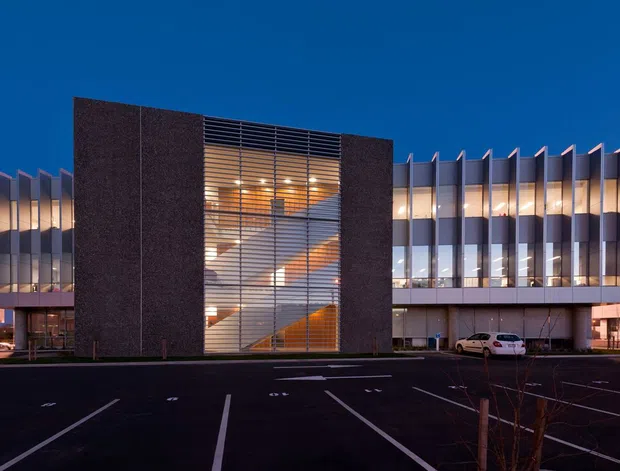 Exterior dusk view of a contemporary multi-storey commercial building. The facade combines a dark solid central volume with a tall glazed stairwell or atrium screened by horizontal louvers, flanked by repetitive floor-to-ceiling glazing and projecting vertical fins. A mostly vacant asphalt parking area occupies the foreground, with warm interior lighting visible through the glazing.