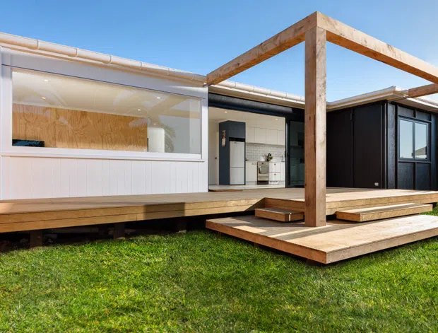 A contemporary single-storey home viewed from the lawn, featuring a raised timber deck and broad timber steps beneath a simple timber pergola. The facade combines light vertical cladding, dark wall panels, large glazed openings, and a partially visible open-plan kitchen inside. The building has a low-pitched roof and a clean, geometric composition.